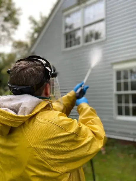 A washer in yellow rain gear soft washing the lap siding of a house