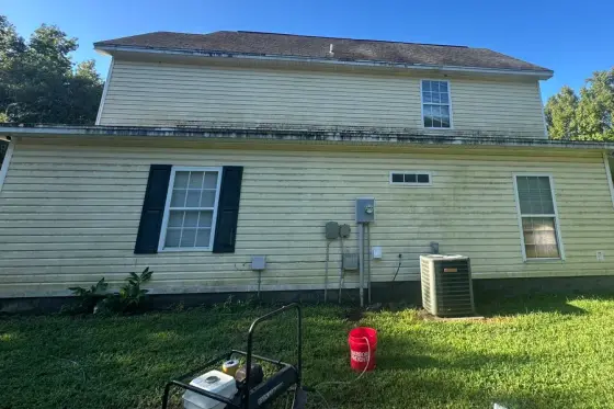 Cream vinyl siding covered in green and black growth, with a pressure washer set up in the yard, Iron Standard job photo